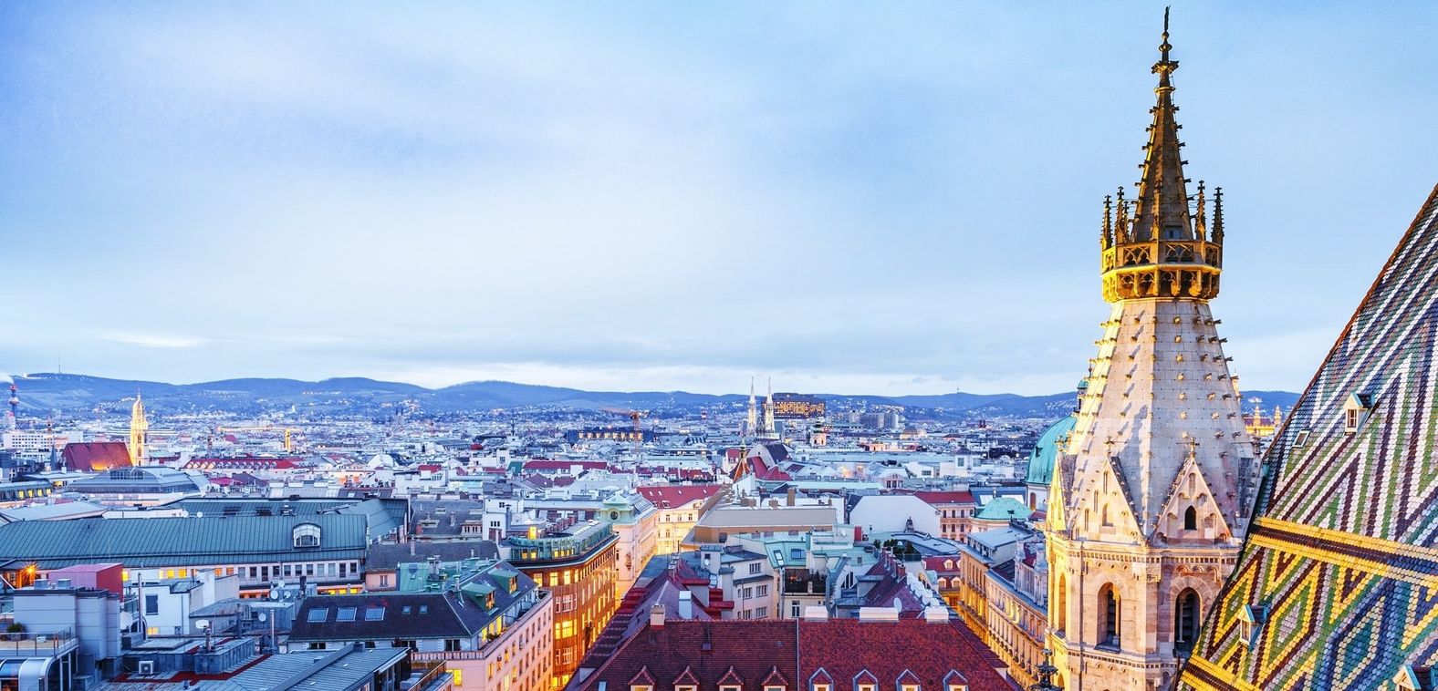 Blick über die Dächer einer Stadt mit einem spitz zulaufenden Turm im Vordergrund und Bergen im Hintergrund unter bewölktem Himmel.